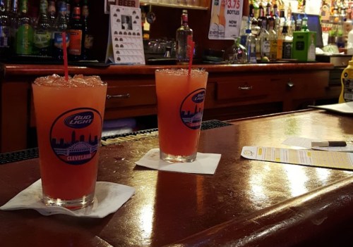 Two colorful cocktails with straws sit on a bar counter, surrounded by various bottles in the background.