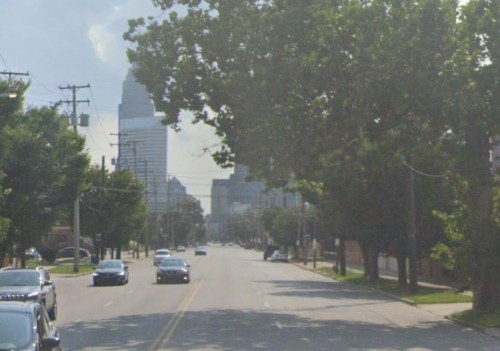A city street lined with trees and cars, leading towards tall buildings in the background under a clear sky.