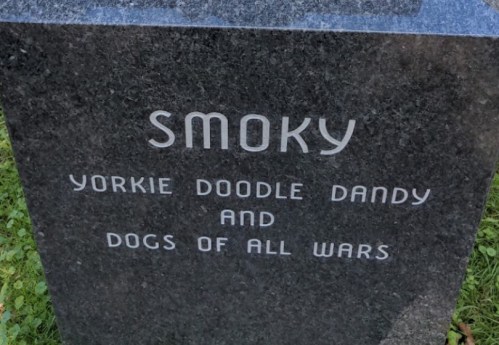 A gravestone engraved with "Smoky, Yorkie Doodle Dandy, and Dogs of All Wars" on a dark stone surface.
