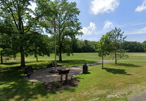 A peaceful park scene with benches, a flagpole, and trees, overlooking a grassy field under a blue sky.