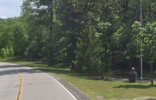 A winding road lined with trees, featuring a bench and a flagpole in a grassy area.