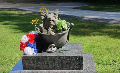 A stone statue of a lion in a bowl, adorned with flowers, sits on a grave in a grassy area.