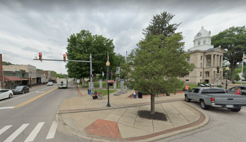 A small town square with a courthouse, trees, and traffic lights, featuring a mix of vehicles and pedestrians.