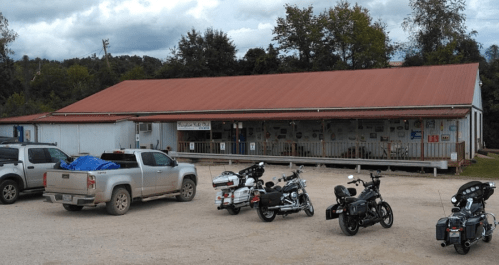 A rustic building with a red metal roof, surrounded by parked motorcycles and a pickup truck on a gravel lot.
