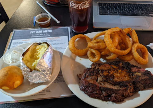 A plate with a baked potato, cornbread, steak, and onion rings, alongside a glass of cola and a menu on the table.