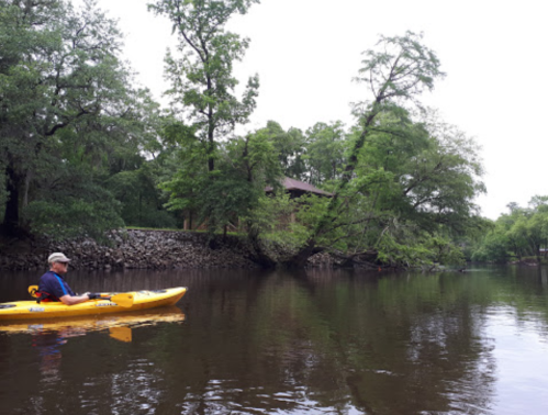 A person in a yellow kayak paddles on a calm river surrounded by lush green trees and a house in the background.