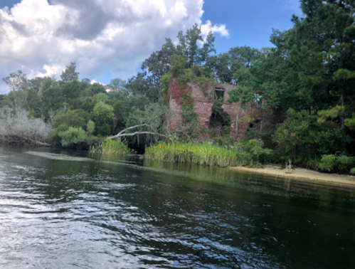 A serene river scene featuring a partially submerged, overgrown brick ruin surrounded by lush greenery and trees.