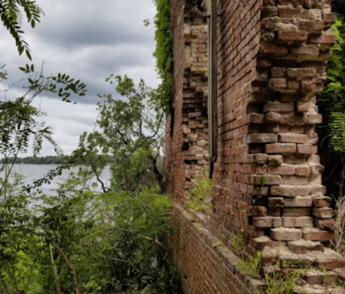 A partially collapsed brick wall covered in greenery, overlooking a body of water under a cloudy sky.
