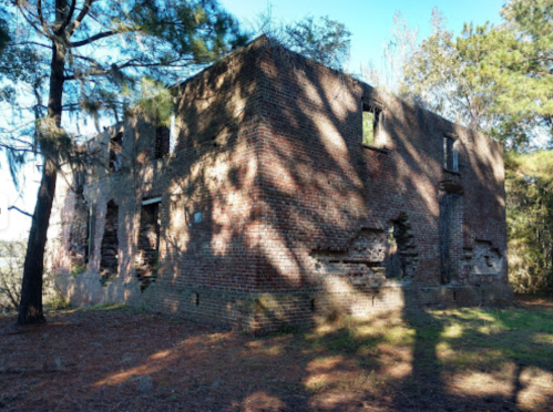 Abandoned brick building surrounded by trees, with visible damage and shadows cast on the ground.