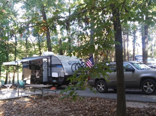 A camper with a bike rack and an American flag parked among trees, with a vehicle nearby and a picnic table in the foreground.
