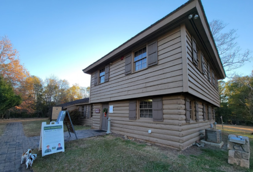 A two-story log cabin with wooden shutters, surrounded by grass and trees, featuring an informational sign and a dog.
