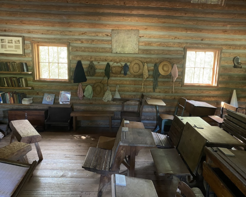 A rustic classroom with wooden desks, bookshelves, and hats hanging on the walls, illuminated by natural light.