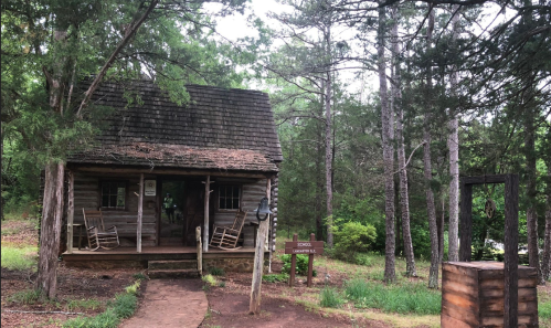 A rustic wooden cabin surrounded by trees, with a porch and rocking chairs, set in a natural landscape.