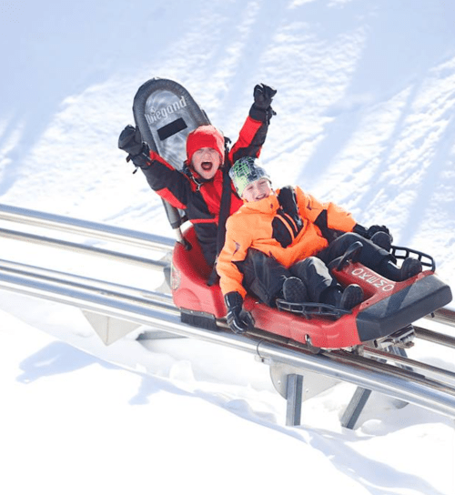 Two children joyfully ride a sled down a snowy track, wearing bright winter gear and smiling with excitement.