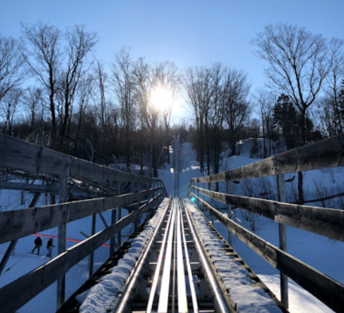 A snowy landscape with a roller coaster track leading downhill, framed by trees and a bright sun in the background.