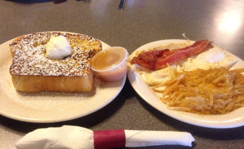 A plate with French toast topped with powdered sugar and a side of eggs, bacon, hash browns, and a small cup of syrup.