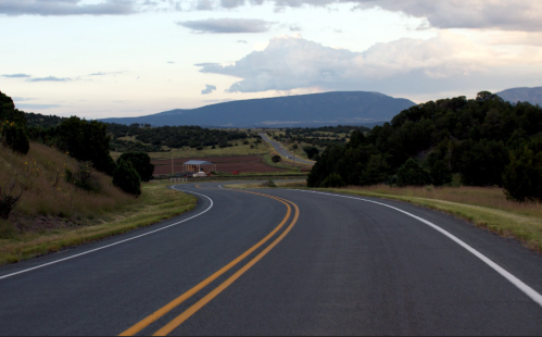 A winding road stretches through a landscape of fields and mountains under a cloudy sky.