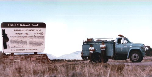 Two people stand beside a vintage blue truck near a sign for Lincoln National Forest, birthplace of Smokey Bear.