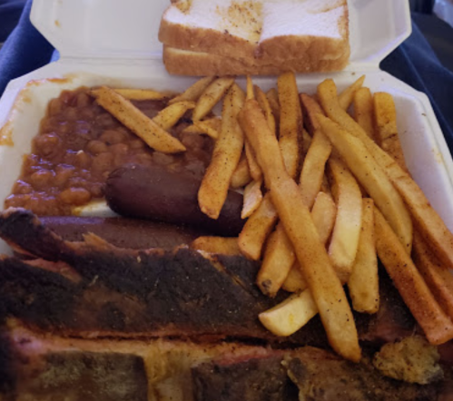 A takeout container with barbecue ribs, baked beans, French fries, a sausage, and slices of bread.
