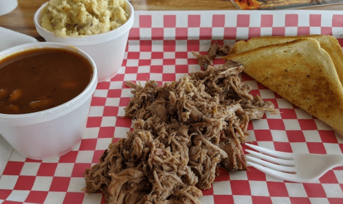 A plate of pulled pork with sides of baked beans, mashed potatoes, and toasted bread on a red and white checkered tray.