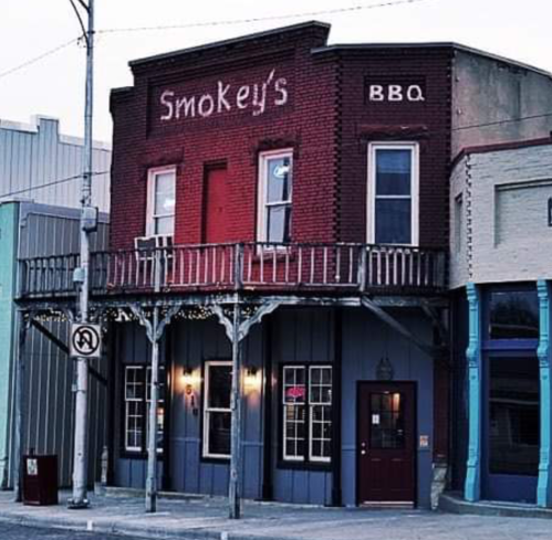 A rustic brick building with a sign reading "Smokey's BBQ," featuring a balcony and colorful storefronts nearby.