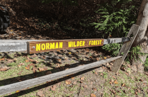 Sign on a wooden fence reading "NORMAN WILDER FOREST" in yellow letters, surrounded by trees and fallen leaves.