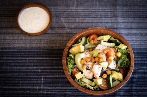 A wooden bowl filled with salad, chicken, avocado, and cherry tomatoes, alongside a small bowl of dressing.