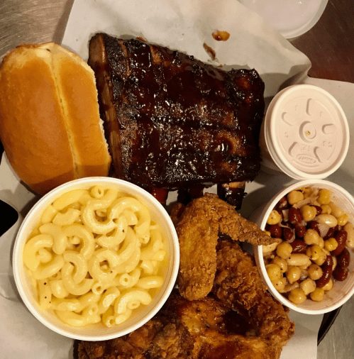 A plate featuring BBQ ribs, fried chicken, macaroni and cheese, and a side of beans, with a bun on the side.