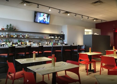A modern restaurant interior featuring red chairs, a bar with bottles, and a TV on the wall.