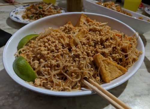 A bowl of stir-fried noodles topped with tofu, crushed peanuts, and lime wedges, served with chopsticks.