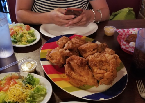 A colorful plate of fried chicken sits on a table with salads and a person using a phone in the background.