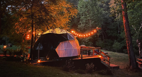 A geodesic dome illuminated by string lights, surrounded by trees in a serene, wooded setting at dusk.