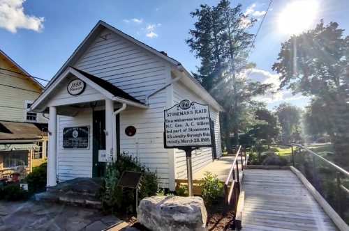 Historic building with a sign about Stoneman's Raid, surrounded by trees and a wooden bridge, under a bright sky.