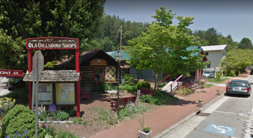 A quaint shopping area with rustic buildings, greenery, and a sign for Old Dillsboro Shops along a quiet street.