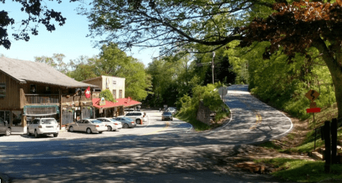 A scenic view of a small town intersection with shops, parked cars, and a winding road surrounded by greenery.