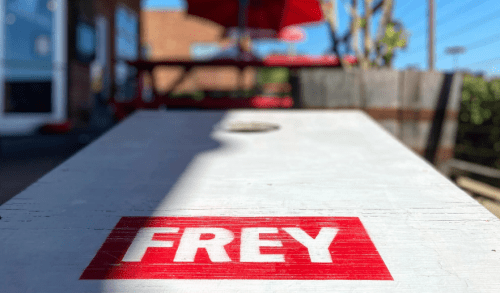 Close-up of a wooden table with the word "FREY" printed in bold red letters, set against a sunny outdoor background.