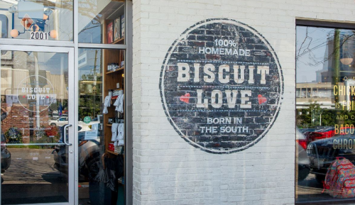 Sign on a brick wall reads "100% Homemade Biscuit Love, Born in the South," outside a restaurant entrance.