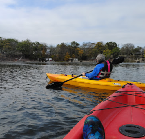 A person in a blue jacket paddles a yellow kayak on a calm lake, with a red kayak in the foreground.