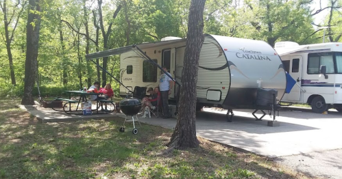 A camper parked in a wooded area, with people sitting at a picnic table and a grill nearby.