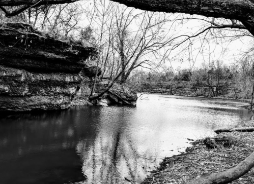 A serene black-and-white landscape featuring a calm river surrounded by rocky banks and bare trees.