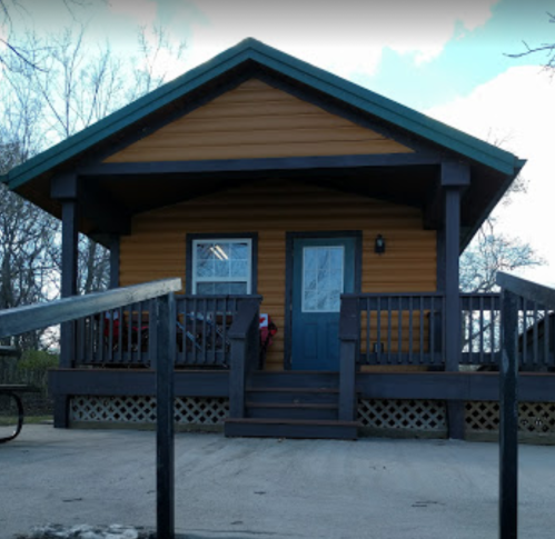 A small, orange and brown house with a porch and steps, surrounded by trees and a clear sky.