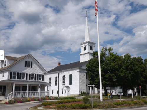 A white church with a tall steeple beside a large tree and a flagpole, set against a cloudy blue sky.