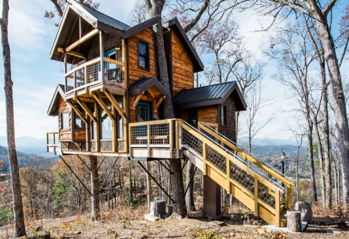 A charming treehouse with wooden exterior, multiple levels, and a ramp, nestled among trees with a scenic view.