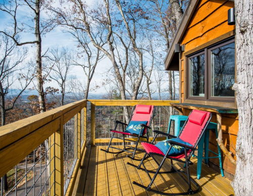 A cozy wooden deck with two red chairs overlooking a forested landscape and blue sky.
