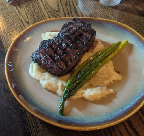 A grilled steak on a bed of creamy mashed potatoes, garnished with asparagus on a round plate.