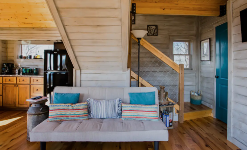Cozy interior of a cabin featuring a sofa, wooden stairs, and a kitchen area with warm lighting and rustic decor.