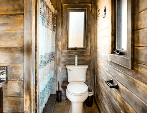A rustic bathroom featuring a wooden wall, a toilet, a shower with a curtain, and a small window.