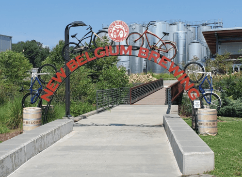 Archway with bicycles and "New Belgium Brewing" sign, leading to a pathway near brewing tanks and greenery.