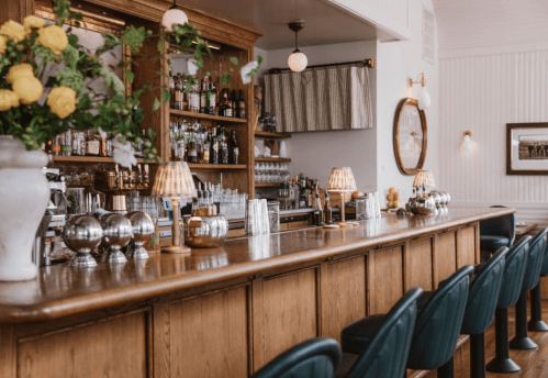 A stylish bar with wooden counters, elegant lamps, and a variety of drinks displayed on shelves in a cozy setting.