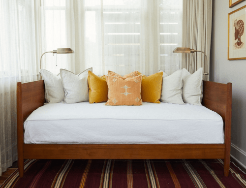 A wooden daybed with white bedding and decorative pillows in a bright, airy room with large windows.